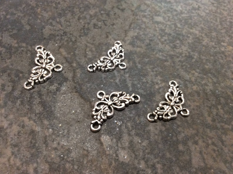May include: Four silver metal corner connectors with a decorative floral design. Each connector has three holes for attaching to other jewelry pieces.
