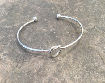 May include: A silver bangle bracelet with a knot design. The bracelet is open and has a simple, elegant style.