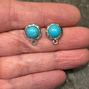 May include: Two silver-toned stud earrings with bright turquoise-colored stones. Each earring has a small loop at the bottom. The earrings are round and set in a silver bezel.