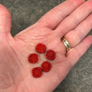 Pu&ograve; includere: Cinque piccoli oggetti rossi a forma di fiore con dettagli scolpiti e caratteri cinesi sono esposti nel palmo aperto di una persona. Gli oggetti hanno una finitura opaca e sono disposti in un gruppo sparso. Lo sfondo &egrave; un grigio neutro screziato.
