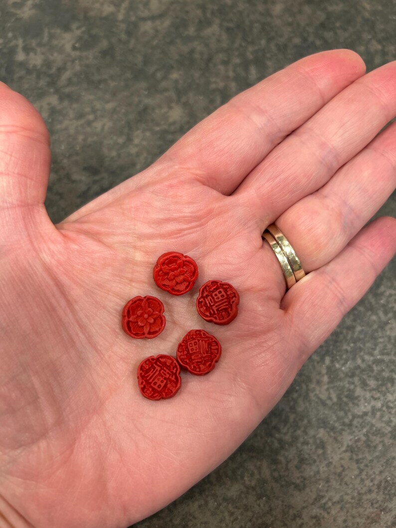 Pu&ograve; includere: Cinque piccoli oggetti rossi a forma di fiore con dettagli scolpiti e caratteri cinesi sono esposti nel palmo aperto di una persona. Gli oggetti hanno una finitura opaca e sono disposti in un gruppo sparso. Lo sfondo &egrave; un grigio neutro screziato.