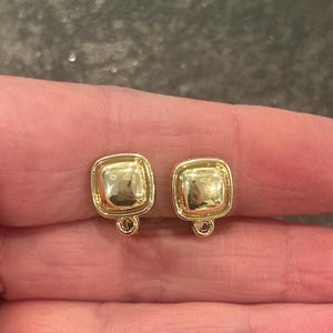 May include: Two gold-colored square stud earrings. Each earring has a rounded square shape with a raised center and a small loop at the bottom. The earrings are held between fingers.
