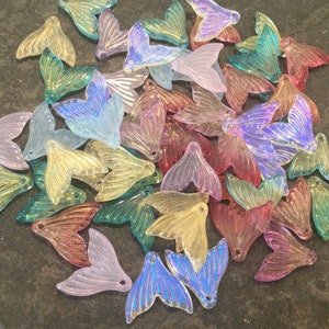 May include: A pile of iridescent mermaid tail charms in various colors, including pink, blue, green, and yellow. The charms are made of clear plastic and have a shiny, glittery finish.
