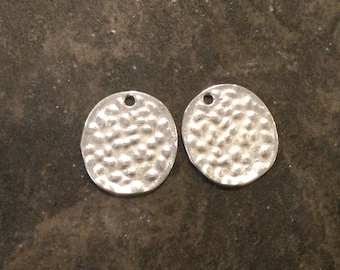 Può includere: Due ciondoli ovali in metallo color argento con una texture martellata e un piccolo foro nella parte superiore.