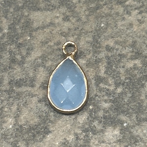 May include: A teardrop-shaped pendant with a light blue gemstone center and a gold-colored metal frame. The pendant has a small loop at the top for attaching to a chain or other jewelry. The gemstone has a faceted design.