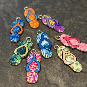 May include: A collection of colorful flip-flop charms with various tropical patterns. Each charm features a gold-colored border and a loop for attaching to jewelry or other crafts. The flip-flops have designs like floral, leaf, and geometric patterns.