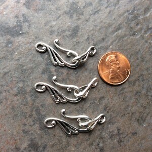May include: Three silver-colored decorative clasps with an ornate, flowing design. Each clasp features a curved hook and a decorative element resembling a stylized wing. A penny is included for size reference.