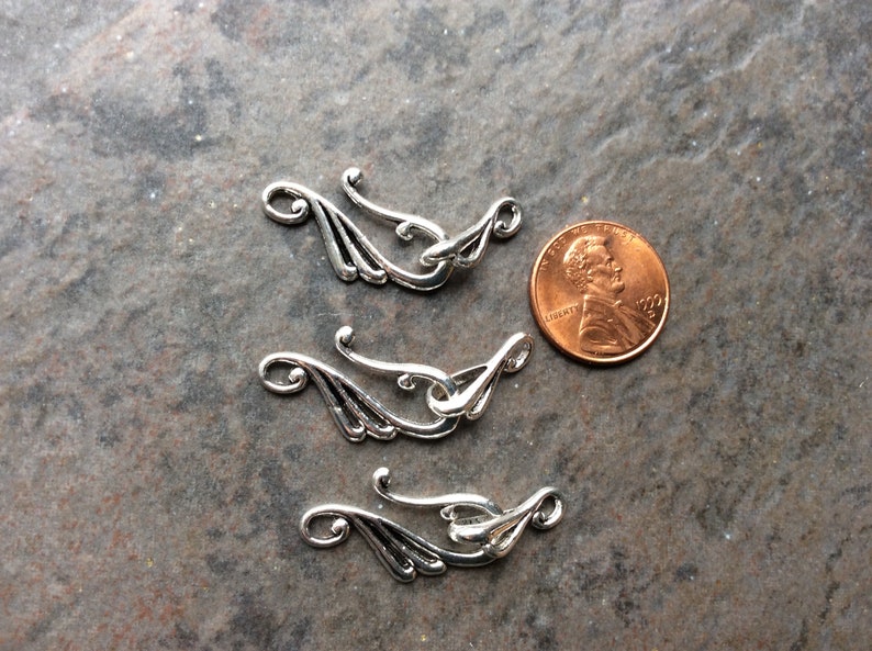 May include: Three silver-colored decorative clasps with an ornate, flowing design. Each clasp features a curved hook and a decorative element resembling a stylized wing. A penny is included for size reference.