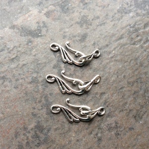May include: Three silver-colored clasps with an ornate, flowing design. Each clasp features a curved hook and a decorative element resembling stylized leaves or wings. The clasps are arranged on a textured, gray surface.