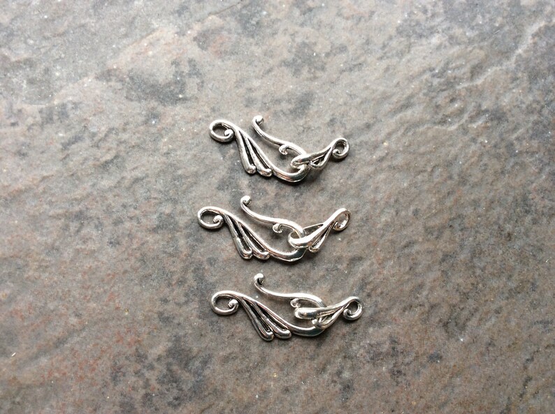 May include: Three silver-colored clasps with an ornate, flowing design. Each clasp features a curved hook and a decorative element resembling stylized leaves or wings. The clasps are arranged on a textured, gray surface.