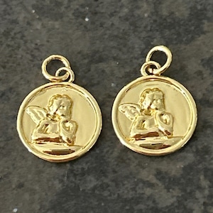 May include: Two gold-colored round pendants. Each pendant features a detailed relief of an angel within a circular frame. The pendants have a small loop at the top for attaching to a chain or other jewelry component.
