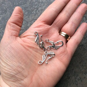 May include: Several small, silver-colored metal jewelry components with a decorative, flowing design are displayed in an open hand. The components have a curved shape with stylized leaf-like details. A gold wedding band is visible on a finger.