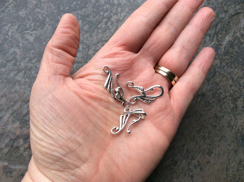 May include: Several small, silver-colored metal jewelry components with a decorative, flowing design are displayed in an open hand. The components have a curved shape with stylized leaf-like details. A gold wedding band is visible on a finger.