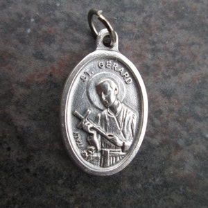May include: Silver oval pendant with a depiction of Saint Gerard Majella holding a cross and a skull. The pendant has the words "St. Gerard" inscribed on it.