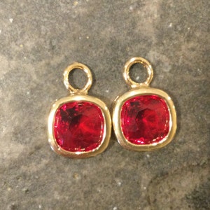 May include: Two gold charms with red square-shaped gemstones. Each charm has a small loop at the top.