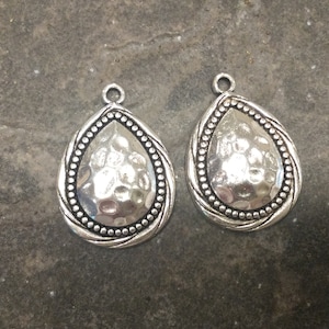 May include: Two silver teardrop-shaped pendant charms with a hammered texture and a beaded border.
