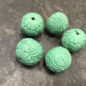 May include: Five round, light green beads with carved floral and geometric patterns. Each bead has a hole drilled through the center. The beads are arranged in a circular pattern on a textured, gray surface. These beads are suitable for jewelry making.
