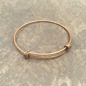 May include: A simple copper expandable bangle bracelet. The bracelet is made of a single piece of wire that is bent into a circle and then joined at the ends with a small clasp.