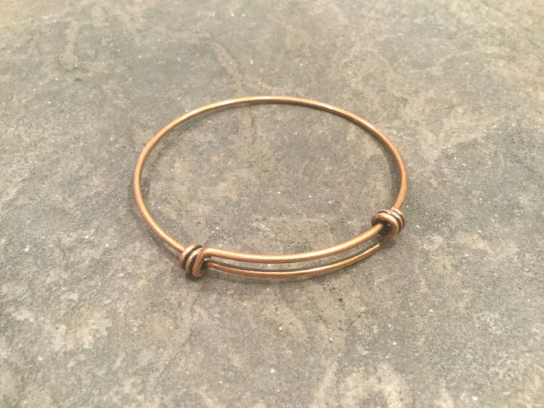 May include: A simple copper expandable bangle bracelet. The bracelet is made of a single piece of wire that is bent into a circle and then joined at the ends with a small clasp.