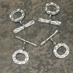 May include: Four silver-colored toggle clasps. Each clasp features a textured, circular ring connected to a textured, rectangular bar. The clasps are designed for jewelry making or craft projects. The clasps are displayed on a textured, gray surface.