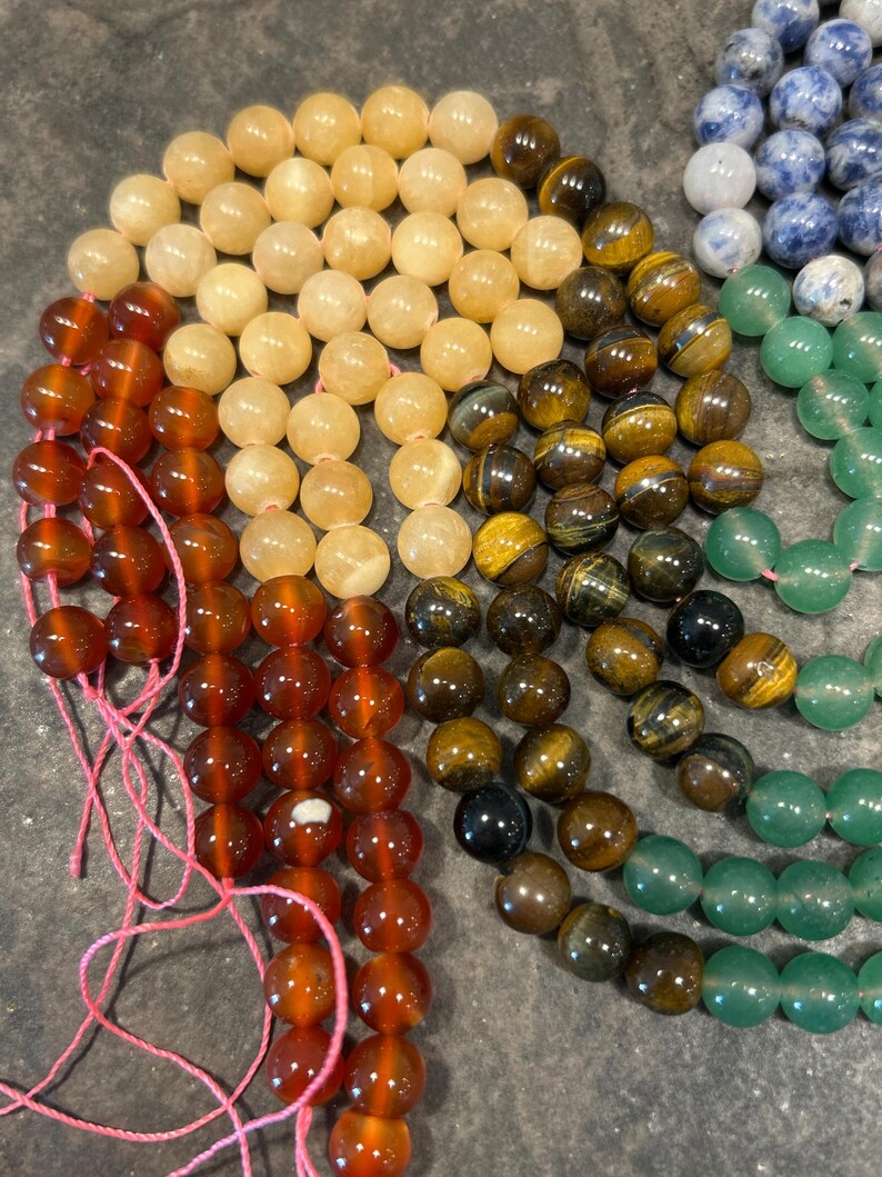 May include: A rainbow of gemstone beads, including carnelian, citrine, tiger's eye, aventurine, and sodalite. The beads are strung on pink thread and are ready to be made into jewelry.