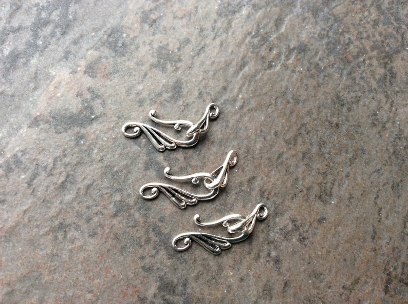 May include: Close-up of four silver-colored metal jewelry findings. Each finding has a curved, wing-like design with a loop at one end and a decorative swirl. The findings are arranged on a textured, gray surface.