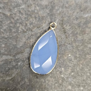 May include: A teardrop-shaped pendant with a faceted, light blue gemstone. The gemstone is set in a gold-colored metal frame and has a small loop at the top for attaching to a chain or other jewelry component. The pendant is displayed on a textured gray surface.