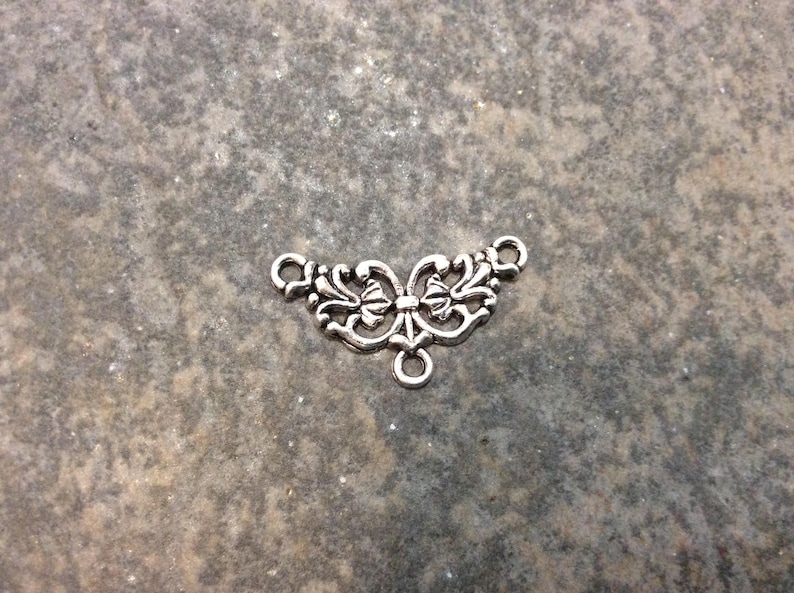 May include: Silver-toned metal connector with a decorative butterfly design. The connector has two loops on the sides and a single loop on the bottom.