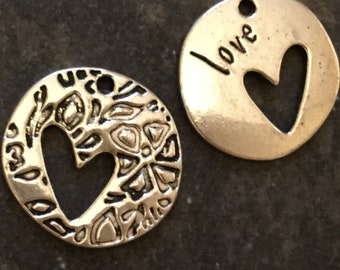 May include: Two silver-toned metal charms. One charm is round with a heart-shaped cutout and floral design. The other charm is round with a heart-shaped cutout and the word "love" engraved. Both charms have a small hole for stringing.