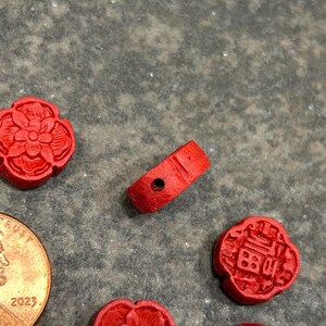 Pu&ograve; includere: Una collezione di piccole perline e ciondoli in legno rosso intagliato. L'immagine include una perlina a forma di anello con un foro e diversi ciondoli a forma di fiore e quadrati. &Egrave; visibile anche una moneta.