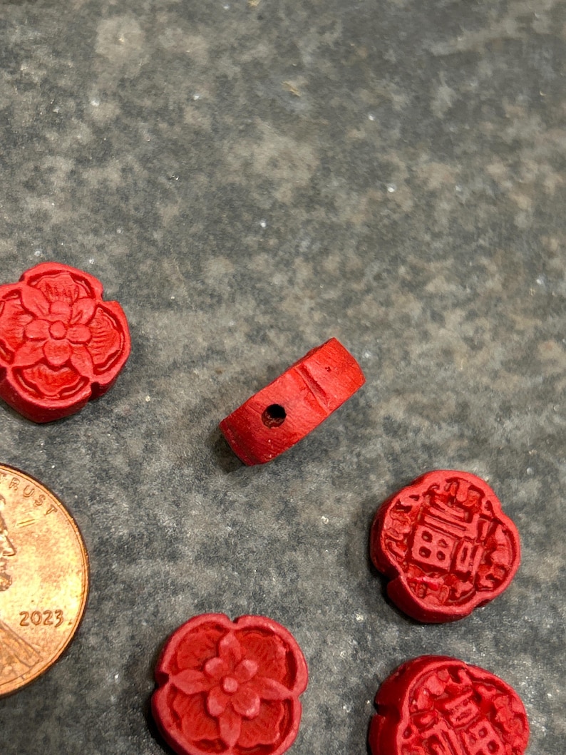 Pu&ograve; includere: Una collezione di piccole perline e ciondoli in legno rosso intagliato. L'immagine include una perlina a forma di anello con un foro e diversi ciondoli a forma di fiore e quadrati. &Egrave; visibile anche una moneta.