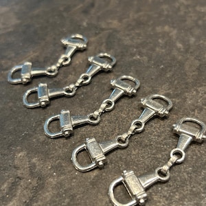 May include: Close-up of five silver-colored horse bit charms. Each charm features a detailed design with a D-ring and a central bit piece, connected by a small chain. The charms are arranged on a dark, textured surface.