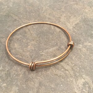 May include: A copper-colored expandable bangle bracelet with a simple design. The bracelet is made of thin wire and has a spring-loaded clasp.