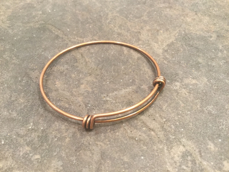 May include: A copper-colored expandable bangle bracelet with a simple design. The bracelet is made of thin wire and has a spring-loaded clasp.