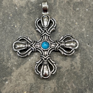 May include: Silver-toned pendant in the shape of a cross with ornate detailing. The cross features a small, round, turquoise-colored stone at its center. The pendant has a loop at the top for a chain. The background is a textured, gray surface.