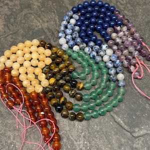 May include: A set of seven strands of gemstone beads, each strand a different color. The colors are, from left to right, orange, brown, green, blue, purple, and pink. The beads are all round and smooth, and they are strung on thin pink cords.