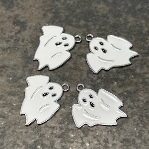 May include: Four white ghost charms with black eyes and silver-colored borders. Each charm has a small loop at the top for attaching to jewelry or other crafts. The charms are on a gray surface.