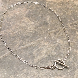 May include: Silver-toned necklace with an oval link chain and a toggle clasp closure. The necklace is displayed on a textured, gray surface. The chain links are elongated and evenly spaced.
