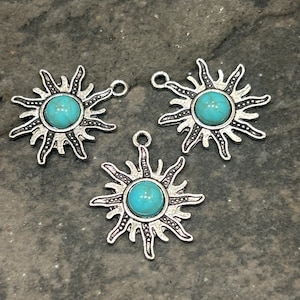 May include: Three silver-toned sunburst charms with turquoise-colored centers. Each charm has a loop at the top for attaching to jewelry. The sunburst design features pointed rays around the central stone.