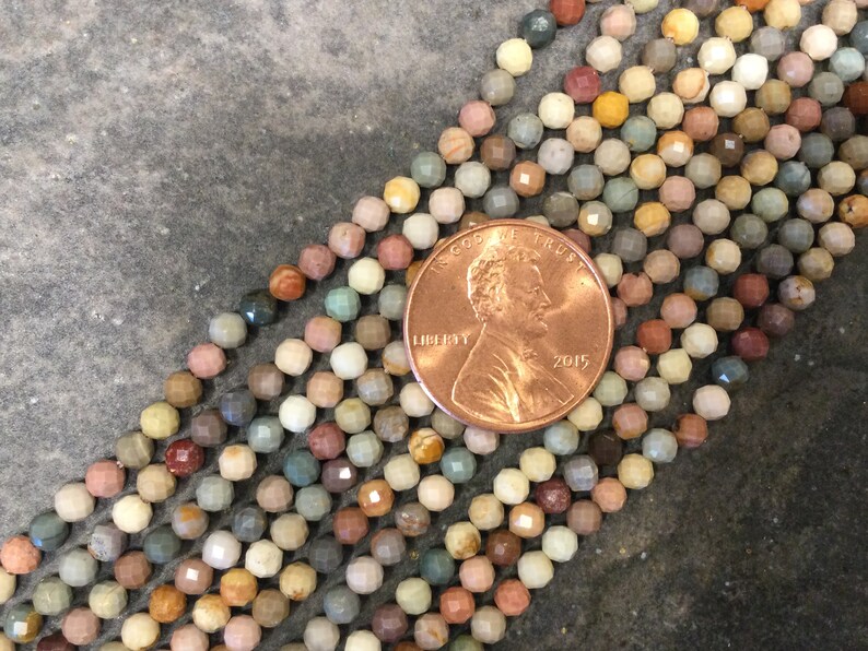 May include: A strand of multi-colored gemstone beads, each bead is faceted and has a unique pattern. The beads are approximately the size of a US penny.