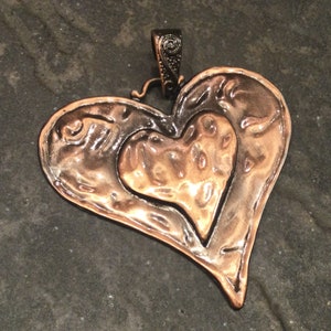 May include: A handcrafted copper pendant in the shape of a heart. The pendant features a textured surface with a smaller heart shape nested inside. The bail is dark metal with a decorative swirl design.