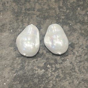 May include: Two iridescent, baroque-shaped pearls. The pearls are white with a shimmering surface, reflecting light. The pearls have an irregular, organic shape, and are set against a textured, gray background.