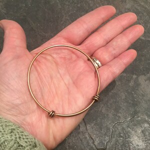 May include: A gold-toned bangle bracelet with two twisted wire accents. The bracelet is shown on a person's hand.