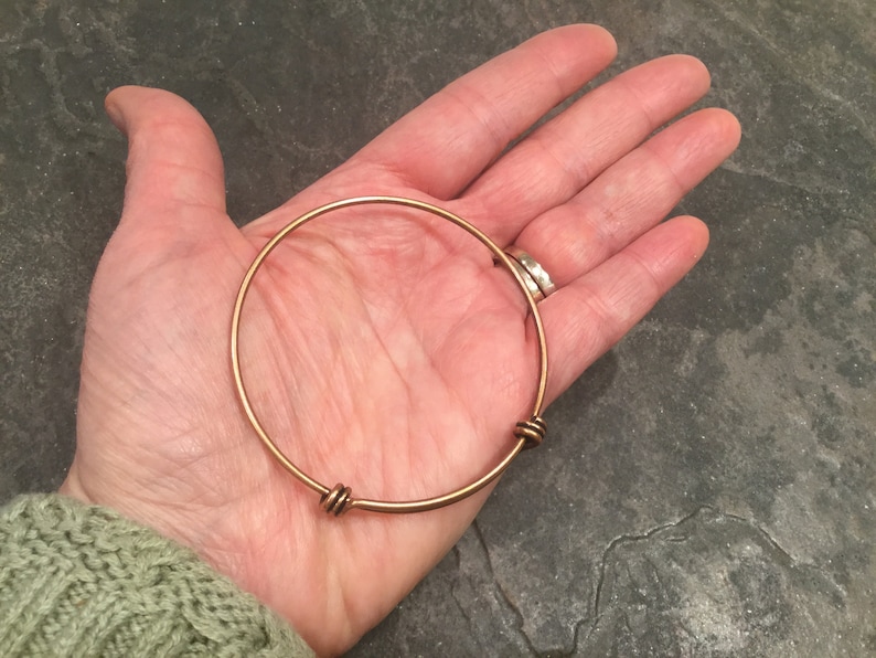 May include: A gold-toned bangle bracelet with two twisted wire accents. The bracelet is shown on a person's hand.