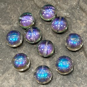 May include: Ten iridescent glass beads are arranged on a surface. Each bead is round with a central groove and displays a spectrum of colours, including blues, purples, and greens. The beads have a galaxy-like appearance with small, white speckles.