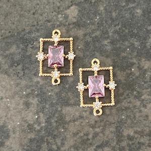 May include: Two gold-colored rectangular pendants with a central pink rectangular gem. Each pendant has a square frame with small, round gold beads and four clear, round gems. The pendants have a small loop at the top for attachment.