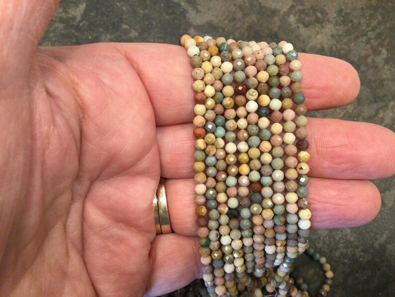 May include: A strand of multi-colored gemstone beads, featuring shades of brown, green, pink, and white. The beads are faceted and have a slightly irregular shape.