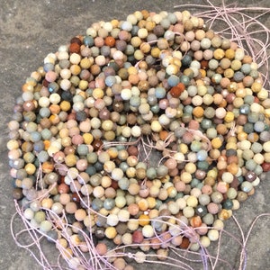 May include: A strand of multi-colored gemstone beads. The beads are faceted and have a rough, natural texture. The beads are strung on a thin, pink thread.