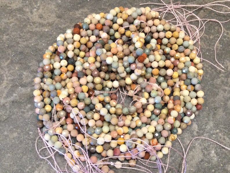 May include: A strand of multi-colored gemstone beads. The beads are faceted and have a rough, natural texture. The beads are strung on a thin, pink thread.