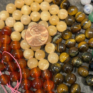 May include: A close-up of three strands of gemstone beads. The top strand is made of light yellow beads, the middle strand is made of brown and gold tiger's eye beads, and the bottom strand is made of reddish-brown carnelian beads. A US penny is included for scale.